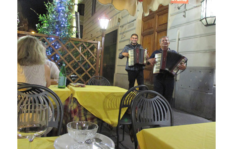 Photo is taken from a small table on the sidewalk, showing two men playing accordions and smiling broadly.